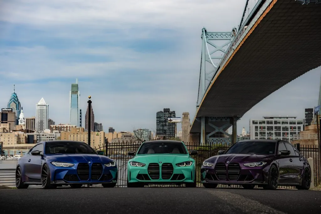 A blue BMW G82 M4 and teal and purple BMW G80 M3s parked under the Ben Franklin Bridge