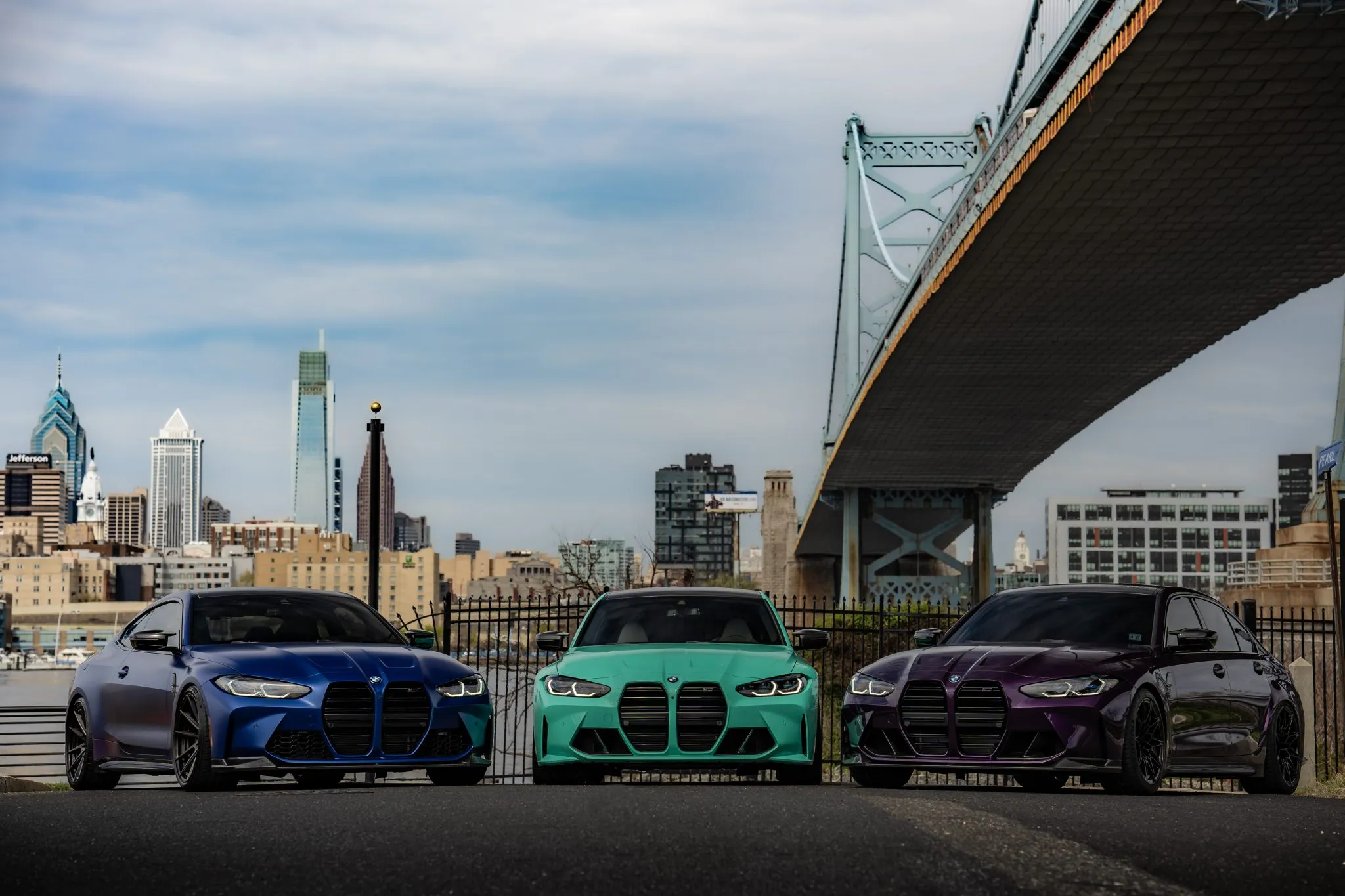A blue BMW M4 and teal and purple BMW M3s under the Ben Franklin Bridge, with the Philadelphia skyline behind